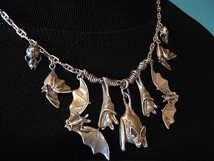 closeup view of a silver necklace featuring multiple batshaped pendants. the necklace has a chain with intricately designed bat charms hanging from it. some bats are depicted in flight, while others hang upside down, mimicking their natural behavior.  there are also small skull charms incorporated into the design. the necklace is displayed on a darkcolored background, which highlights the silver detailing. the overall style is gothic or darkthemed.