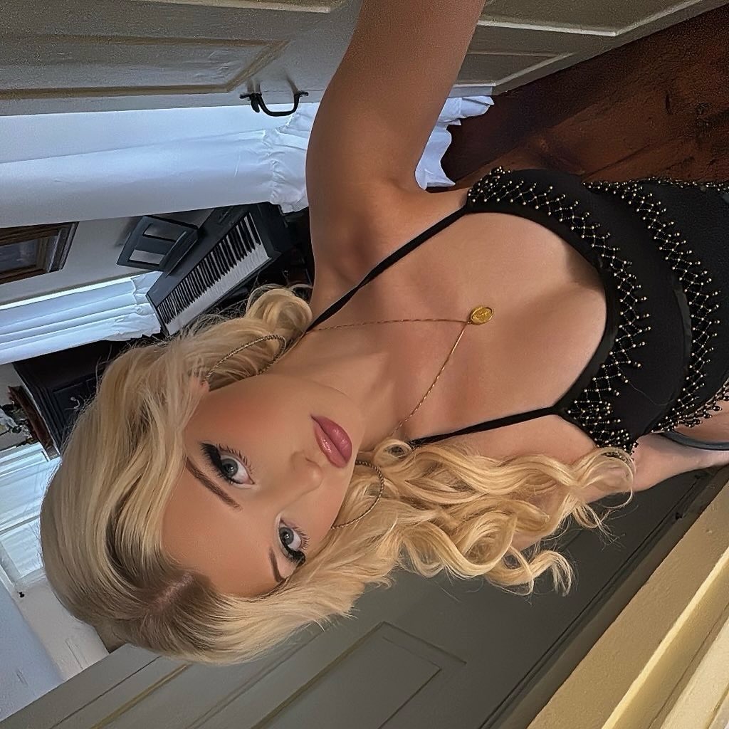 a woman with blonde hair is wearing a black dress and jewelry, including a necklace. she is posing for a selfie. a piano is visible in the background.