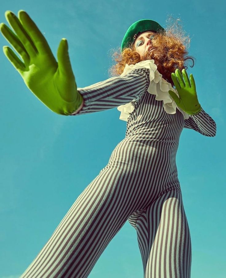 a woman wearing a green hat and striped jumpsuit with green gloves is posing with her arms stretched out. the background is a clear blue sky.