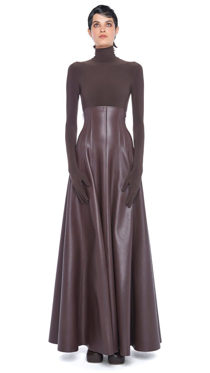 a woman in a long brown leather skirt and matching turtleneck top poses for a studio photoshoot against a white background. she is wearing long brown gloves and a chic updo. the outfit is elegant and formal, with a high waist and a flowing skirt. the model is looking up and slightly to the right, with a confident expression.