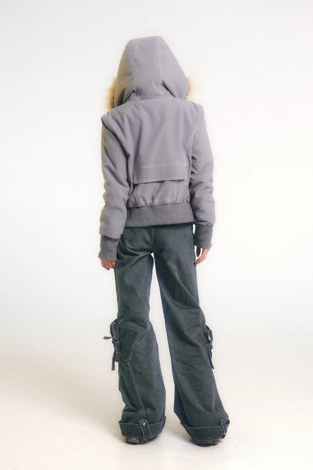 a full shot of a female model standing with her back to the camera, against a white background.  she's wearing a grey cropped hooded jacket with a fur trim on the hood. the jacket is puffy and appears warm. she is wearing dark grey cargo pants with ties at the ankles.  the overall style is casual and winterappropriate.
