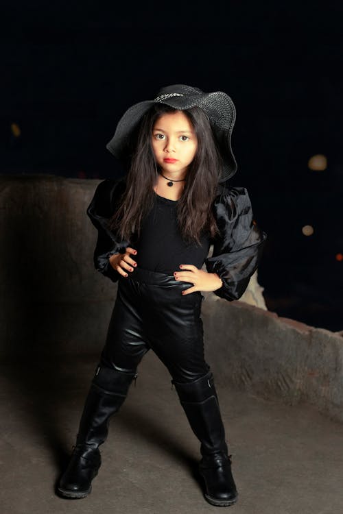 a young girl, possibly a child model, strikes a confident pose in a stylish allblack outfit. she's wearing a widebrimmed black hat, a black longsleeved top with puffed sleeves, black leather pants, and tall black boots. her dark hair is long and straight. the background is dark and urban, suggesting a nighttime setting. the overall aesthetic is edgy and cool. the girl's expression and pose exude confidence and attitude.