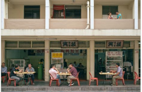 a shot of a restaurant in a busy city. people are seen sitting at the tables in front of the restaurant. the restaurant has a red sign above the door. the sign has writing in a foreign language. the restaurant has a view of a busy street. the restaurant has a lot of windows and a lot of natural light. there is a lot of food and drink available.