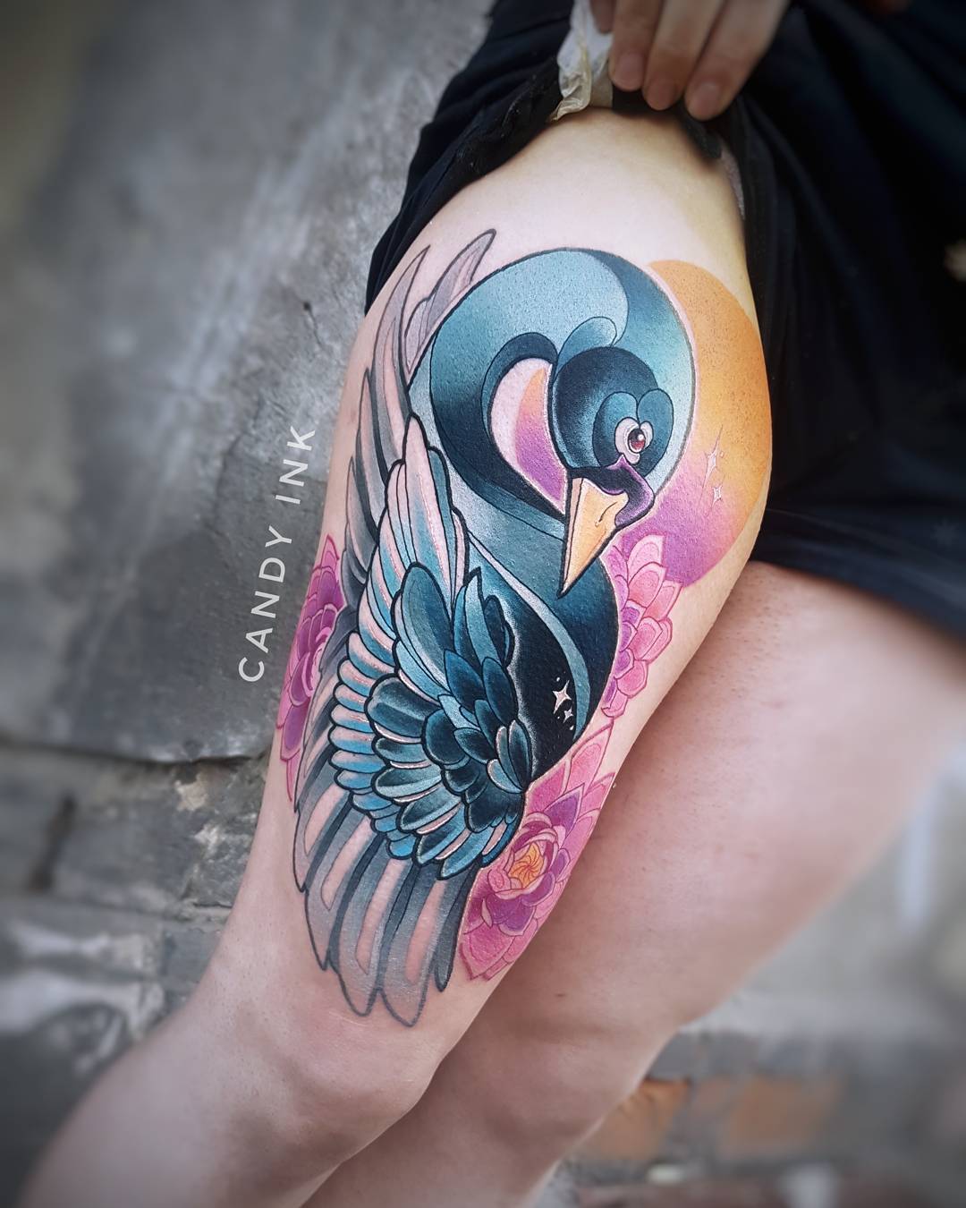 a neotraditional style tattoo of a swan on a person's thigh. the swan is depicted in blue, black, and white, with pink lotus flowers and a yellow sun in the background. the tattoo is done in a painterly style, with smooth shading and vibrant colors. the tattoo artist's signature, "candy ink", is visible in the upper left corner of the image.