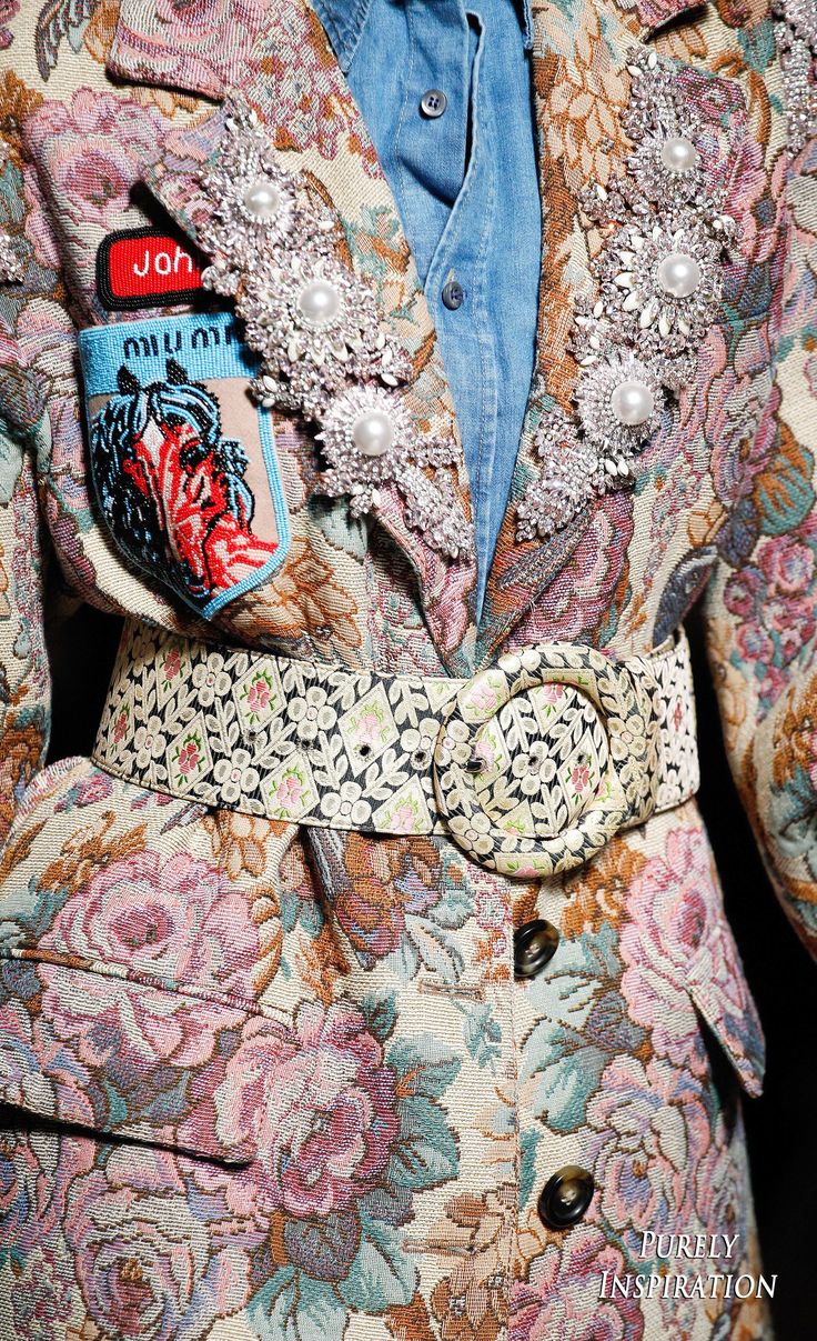 loseup detailed shot of a woman's floral tapestry jacket. the jacket features a patterned belt,  pearl and rhinestone embellishments, and patches with text and images. the jacket is richly textured, displaying a variety of colors and patterns, and appears to be a vintage or highfashion garment. a denim shirt is visible under the jacket.  the overall aesthetic is luxurious and unique.