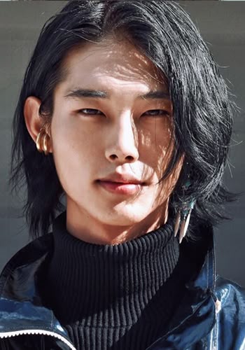 a portrait of a young asian man with long black hair wearing a black turtleneck and a shiny jacket. he is wearing gold hoop earrings and feather earrings. he is looking directly at the camera. the lighting is bright and creates shadows on his face.