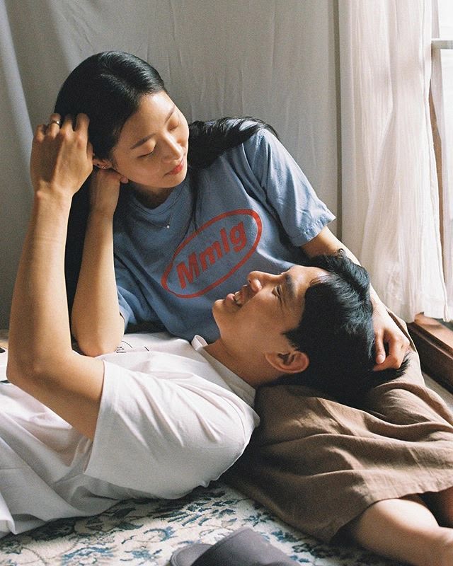 a young asian couple is lying down indoors, gazing at each other with an intimate and relaxed expression. the woman, wearing a blue tshirt with the letters "mmig" in a red oval, is propped up on her arm, looking down at the man. the man, dressed in a white tshirt, is lying with his head resting on a brown duvet or pillow. they appear to be in an apartment or home setting, with a patterned rug beneath them and a white curtain and window providing natural light. the scene conveys a sense of coziness, love, and shared moments between the couple.