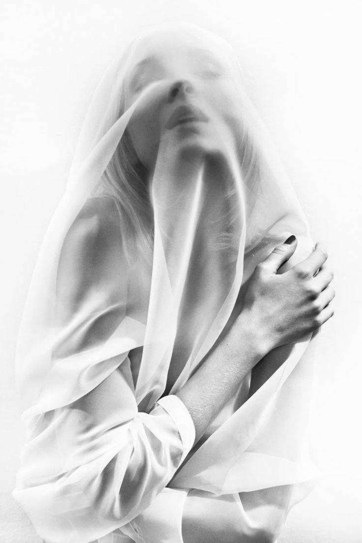 the image is a black and white closeup portrait of a woman partially obscured by a sheer, white fabric draped over her body. the fabric covers most of her face, creating a sense of mystery and leaving only her eyes and some facial features visible.  her arms are visible and she holds the fabric close to her body. the overall feel is artistic, sensual, and minimalist, with a focus on texture, light, and shadow. the monochromatic palette emphasizes the softness and elegance of the scene.