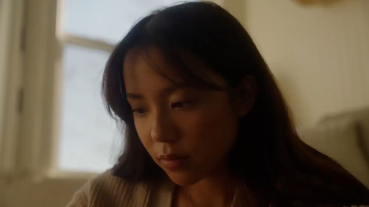 the image shows a young woman with long dark hair and brown eyes. she is wearing a beige sweater and is lit by warm, natural light coming from a window to her left. her expression is calm and slightly pensive, as if she is deep in thought. the overall atmosphere of the image is serene and peaceful.
