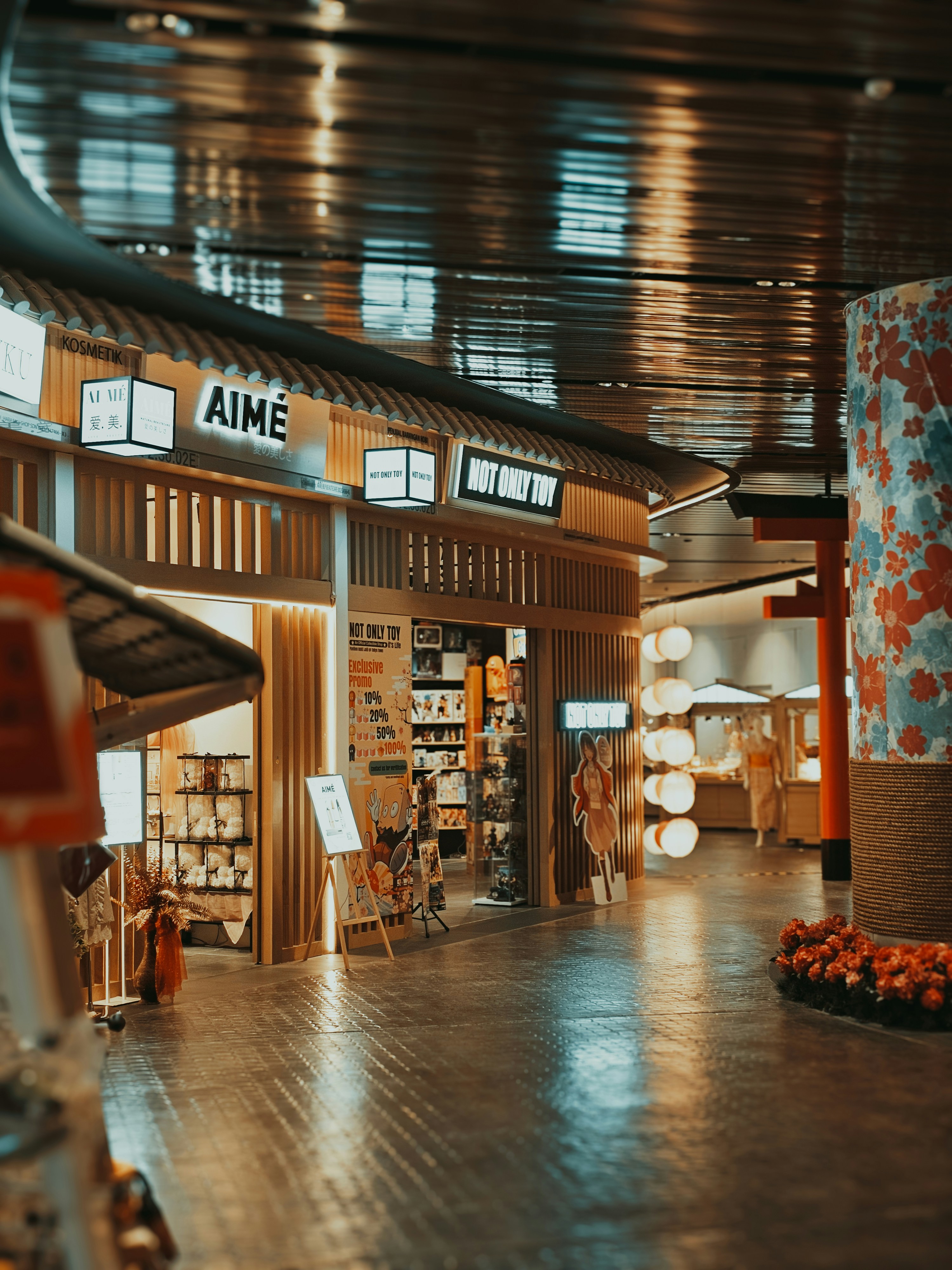this image shows the interior of a mall designed to look like a traditional japanese shopping street. the shops have warm wood paneling and signage with japanese characters. the floors are tiled and glossy, reflecting the artificial lighting overhead. there are very few people in the image, with only one partially obscured individual visible in the distance.  the overall atmosphere is quiet and inviting.