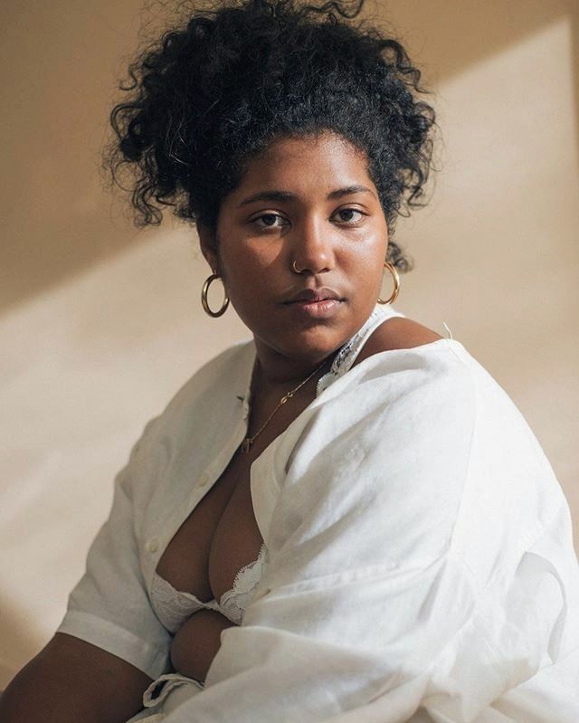 a portrait of a woman with curly dark hair, wearing a white buttondown shirt open to reveal a white lace bra. she has hoop earrings and a nose ring, and a gold necklace. the background is a neutral tan color, and soft light falls across her face and upper body.