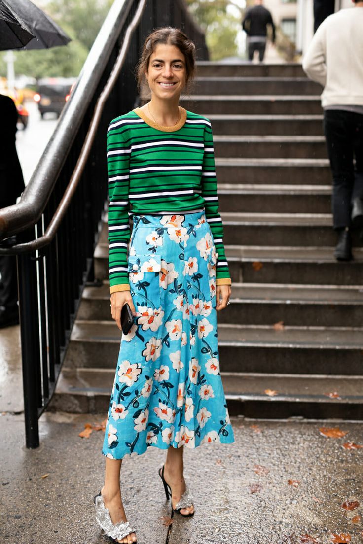 a woman stands on a set of stairs, smiling at the camera. she is wearing a green and white striped longsleeved sweater and a light blue floral midi skirt. she is also wearing silver highheeled sandals. the photo is taken outdoors, in a city setting, likely in autumn due to the weather and the colors of the clothing. the overall aesthetic is stylish and fashionable.