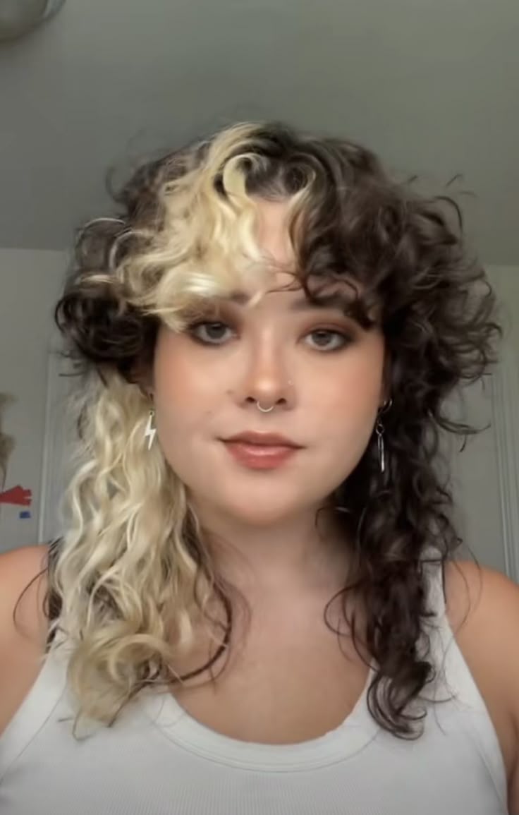 a woman with splitdyed curly hair, one side blonde and the other dark brown, is wearing a white tank top, earrings, and a septum piercing. she has makeup on.