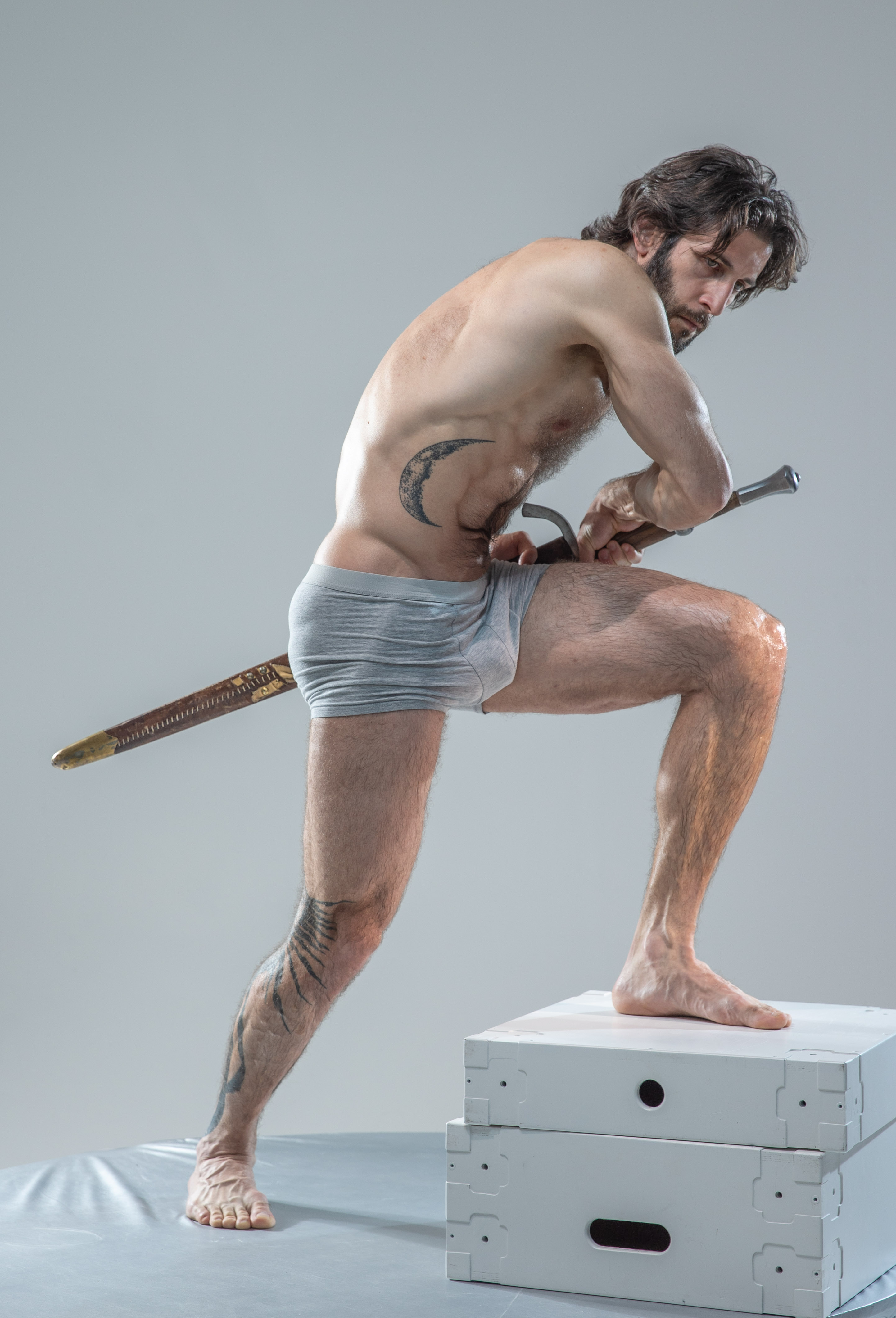 a muscular, barechested man with tattoos stands in a dynamic pose atop a white platform in a studio setting. he holds a sword with a determined expression, his body tensed for action. the image is lit with bright, artificial lighting, emphasizing his physique and the details of his pose.