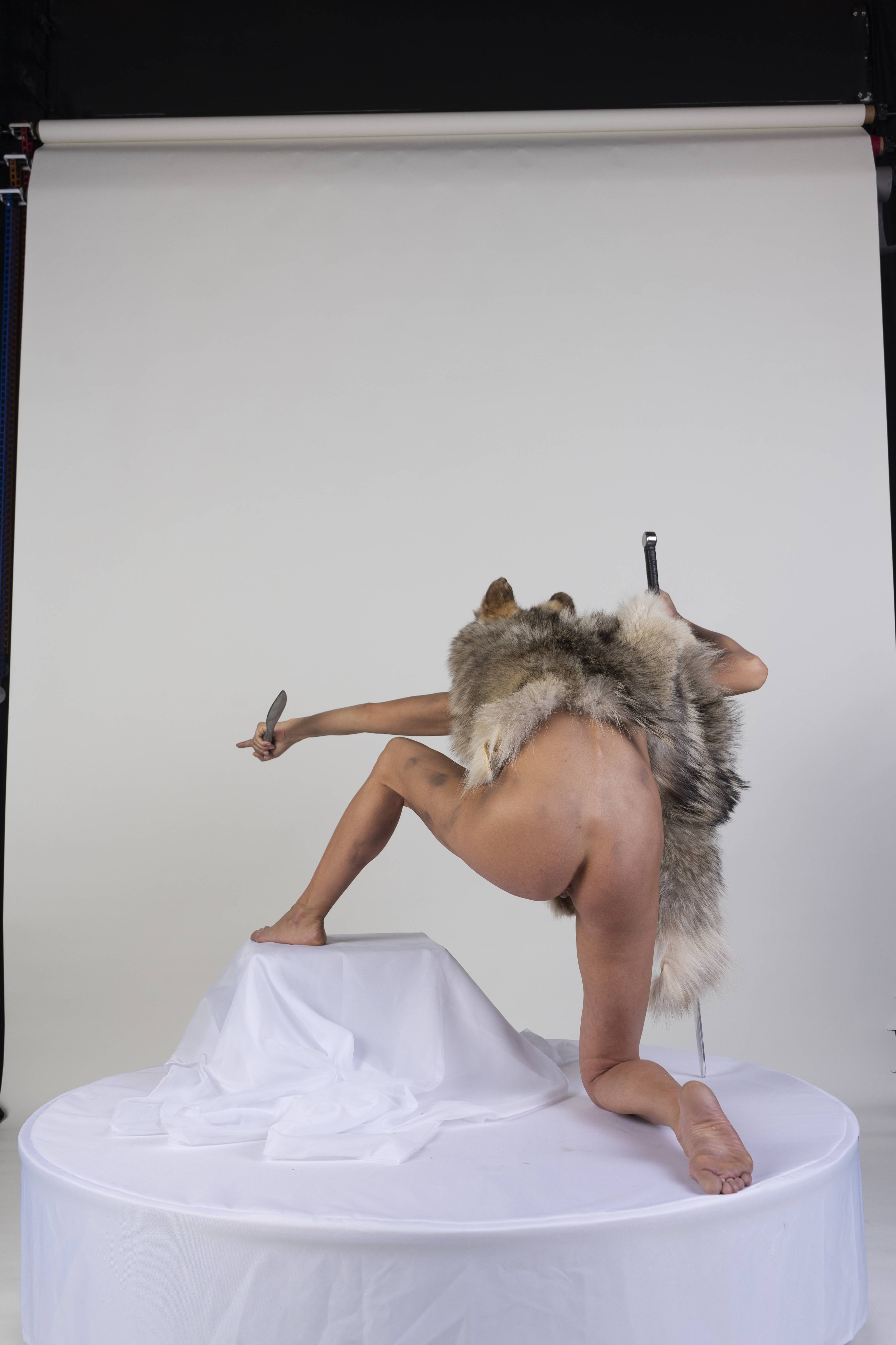 a nude woman is kneeling on a white pedestal in a studio with a white background. she is wearing a fur shawl and holding a knife in her right hand. a sword is attached to her back.