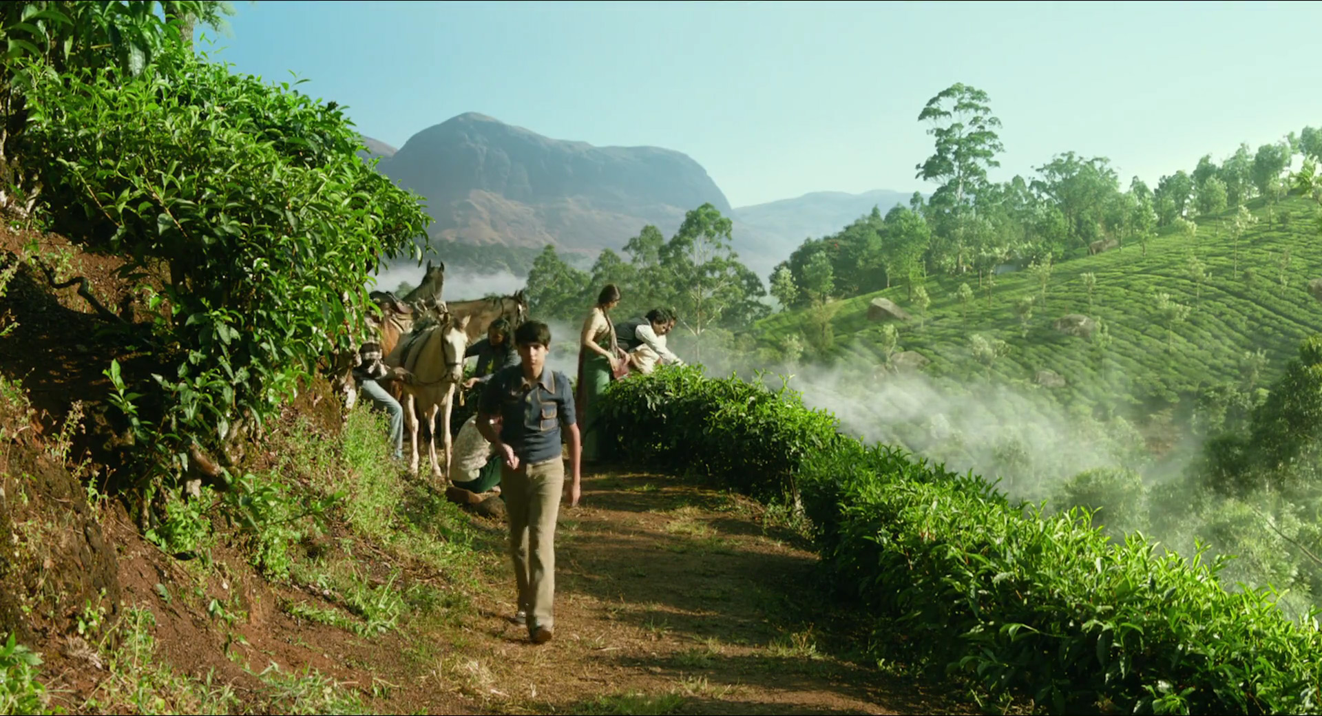 a group of people are walking along a dirt path in a tea plantation. the path is lined with lush green tea plants, and in the background, there are rolling hills and mountains. the sun is shining brightly, casting long shadows on the ground. there are horses standing on the path, and some people are riding them. the overall atmosphere of the image is one of peace and tranquility.