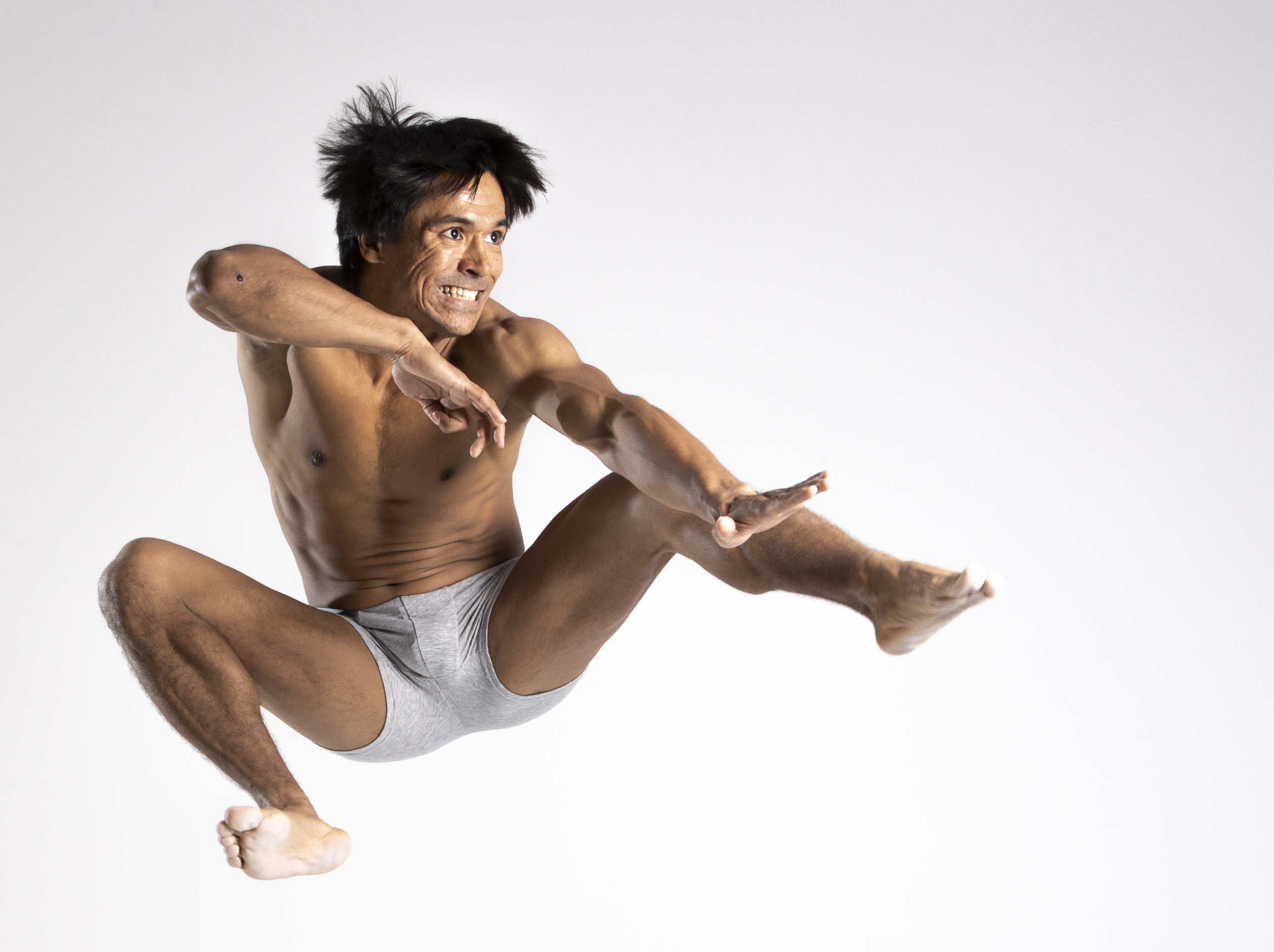 a shirtless man wearing gray underwear is captured midair in a dynamic jumping pose reminiscent of martial arts. his arms are outstretched, and his expression is intense as he appears to be executing a move. the image is a studio shot with a clean white background, emphasizing his muscular physique and athletic prowess. he is barefoot and frozen in action.