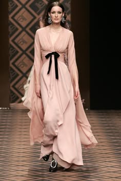 a beautiful woman in a long pink dress, poses for a runway fashion show. she is wearing a black bow, black shoes, and jewelry. her hair is pulled back and she looks elegant and stylish. the dress is flowing and graceful and she is posing with her hand on her hip. the background is a blurry, abstract pattern, suggesting a luxurious backdrop. the image captures the essence of fashion and elegance.
