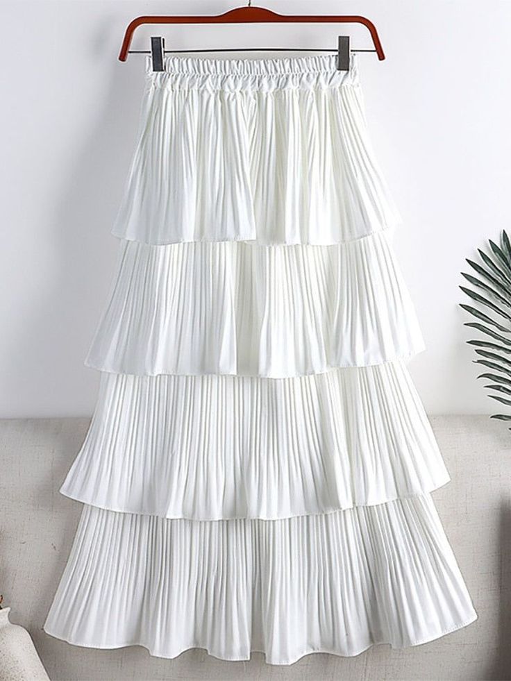 a white pleated maxi skirt with multiple layers is hanging on a wooden hanger. the skirt is made of a soft and flowy chiffon fabric, and it has a delicate and feminine feel.  it is perfect for a summery look, and can be dressed up or down to suit any occasion.