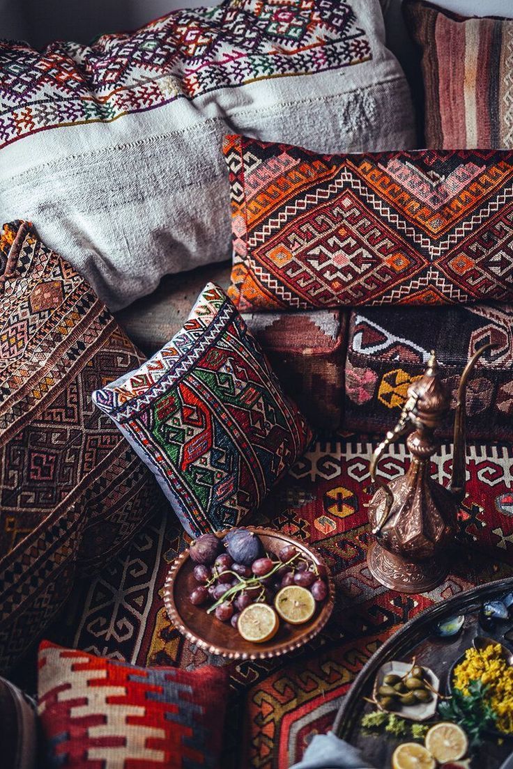 a view of a lavishly decorated oriental setting featuring an abundance of patterned textiles and pillows. a decorative hookah sits prominently on the rug, along with platters of fruit and snacks, including figs, grapes, lemons, and other unidentified delicacies.
