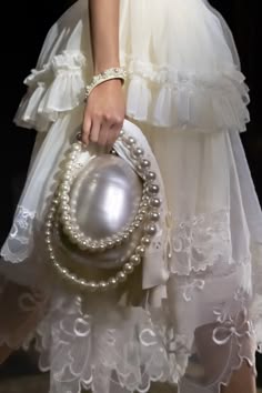 a woman in a white lace dress is holding a pearl and white clutch bag. she has a pearl necklace and bracelet on her wrist. the woman is styled in a vintage fashion, with a dress that is flowing and delicate. the dress has a lot of lace detail and a ruffled hemline. the pearl clutch and necklace are the perfect accessories to complete the look. she appears to be looking at the camera, but her face is not visible.