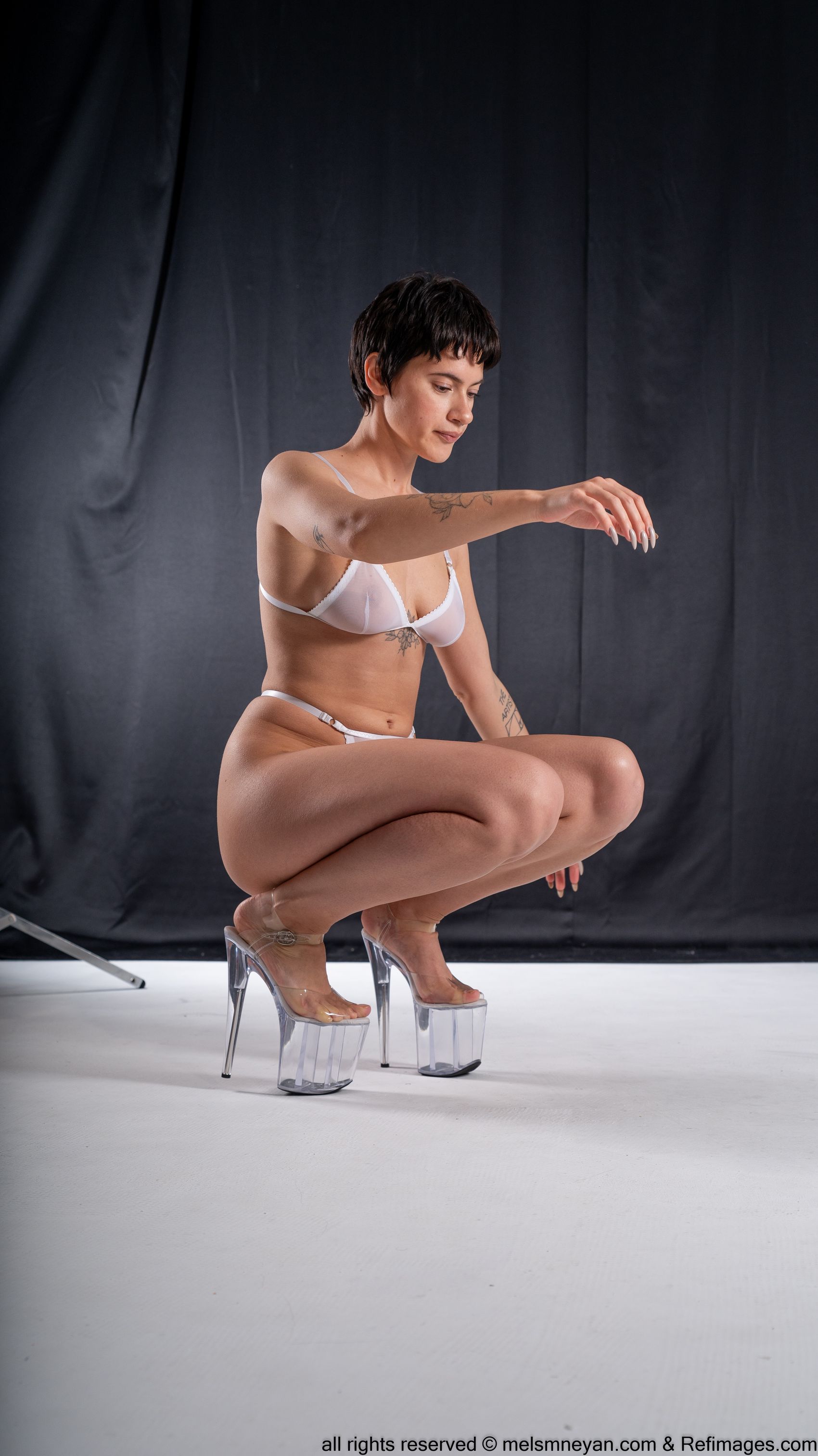 a woman with short, dark hair is squatting on a white surface in front of a dark background. she is wearing white lingerie and clear, platform heels. she has tattoos on her arms and feet, and long nails. she is looking down and reaching out with her right hand.