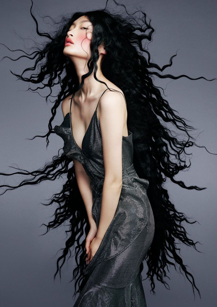 loseup shot of a fashion model with extremely long, dark, curly hair that flows dramatically around her. she's wearing a shimmering gray, formfitting dress with a deep vneckline and ruffled detailing. the model is positioned in a graceful pose, her gaze directed upward. the background is a muted gray, which emphasizes the contrast of the model's dark hair and attire. the overall impression is one of elegance and sophistication, highlighted by the intricate styling of her hair and the simplicity of her dress. the image appears to be a professional fashion photograph or editorial shot.