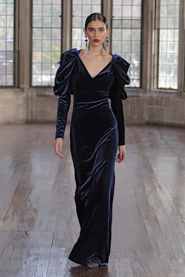 a fashion model walks down a wooden runway, showcasing a dark blue velvet gown. the dress features long sleeves with puffed shoulders and a vneckline. the model's makeup is simple and elegant, with a red lip. the background includes large windows showing a view of what seems to be a stone building. the overall mood is sophisticated and elegant.