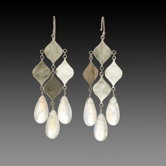 a pair of chandelier earrings made from sterling silver and moonstone gemstones dangle against a dark gray background. the earrings feature multiple teardropshaped moonstones suspended from hammered silver diamond shapes.