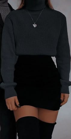 a woman wearing a black sweater, a black skirt, and black tights stands with her legs crossed. she has a heart necklace around her neck.