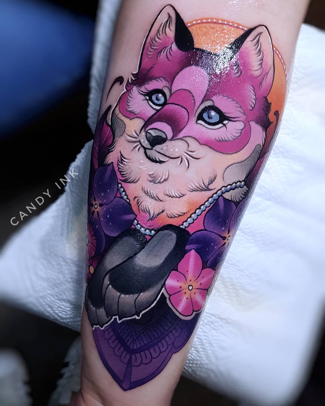 a closeup photograph showcasing a pink fox tattoo on an individual's arm. the fox is stylized with large blue eyes, surrounded by purple flowers, and rendered in shades of pink and white.  the artistry exhibits clean lines and vibrant colors.  the photo is likely taken within a tattoo parlor given the environment and lighting.