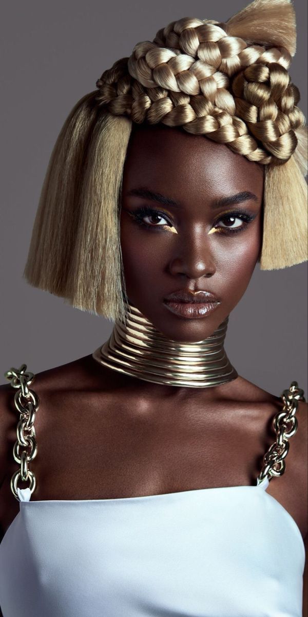 a black woman with blonde, braided hair is wearing a thick gold necklace and a white top with gold chain straps. she is looking directly at the camera. the portrait is taken in a studio setting.