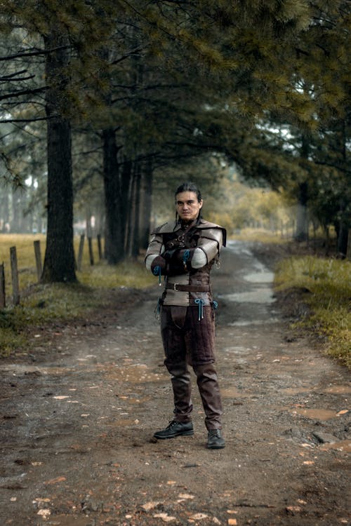 a full shot of a man dressed in a medievalstyle costume, possibly cosplay, standing on a dirt road in a forest. he is wearing brown leather armor and pants, and has his arms crossed. he appears serious and is looking directly at the camera. the setting is a forest with tall trees lining a path. the overall mood is dark and mysterious, suggestive of a fantasy or historical setting.