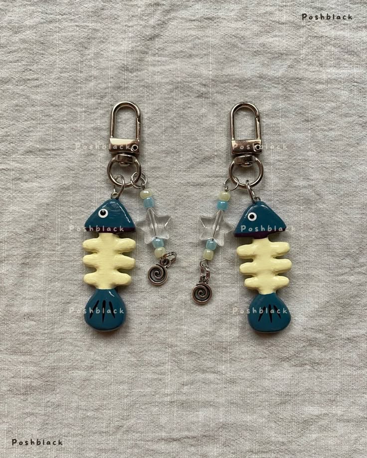 two fishbone keychains with blue and yellow fishbone charms, clear and blue beads, silver star charms, and spiral charms are presented on a textured light grey background. the keychains are attached to metal hardware. the image also includes a watermark "poshblack" in multiple locations.