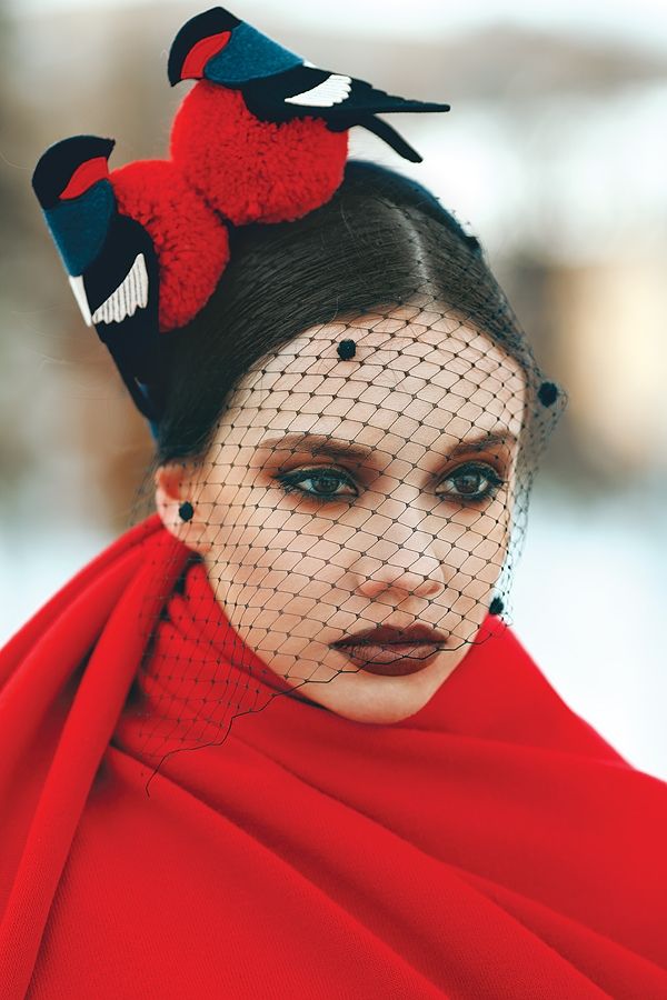 a portrait of a woman wearing a red scarf and a hat decorated with bird figures. she also has a black dotted veil covering her face.