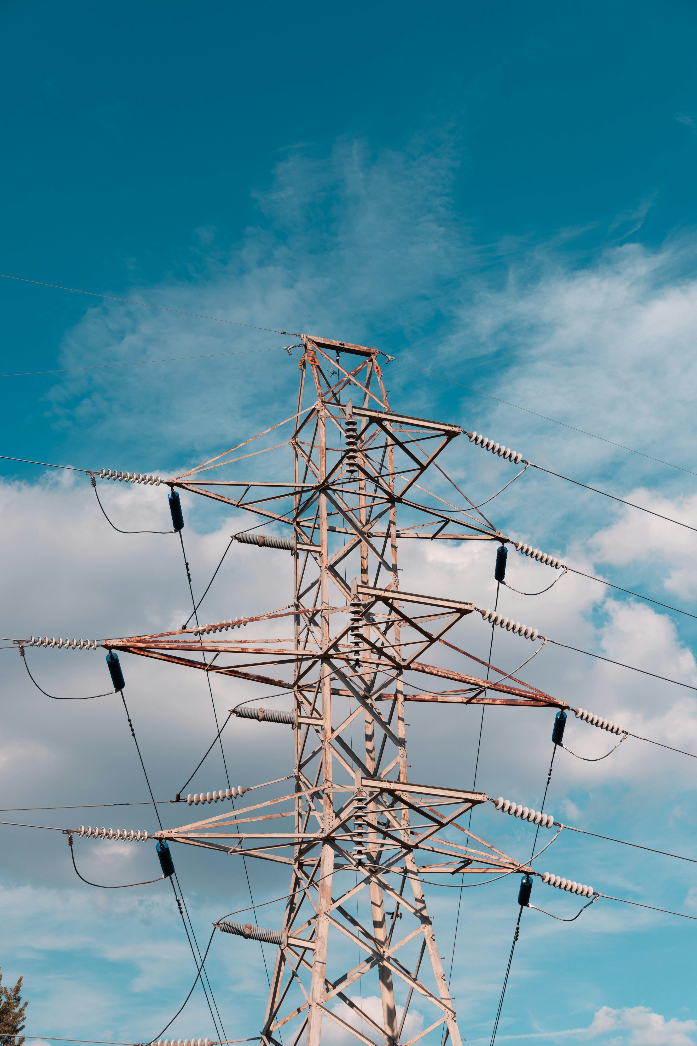 the image shows a tall electricity pylon against a bright blue sky with scattered white clouds. the pylon is made of a lattice of metal beams and is supported by wires that extend in different directions. the image is taken from a low angle, making the pylon appear imposing against the vastness of the sky.
