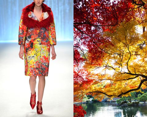 a woman wearing a red furtrimmed coat with colorful floral print is standing on a white runway. the background is a blurry view of a lake and autumnal trees in a park. the trees have a mix of yellow, orange, and red leaves.  the woman is wearing red stiletto heels and has a red leather bag.  the overall image is one of beauty, fashion, and the changing seasons.