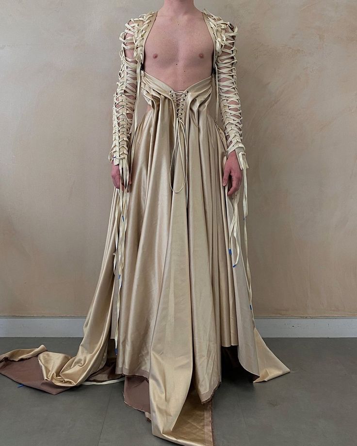 a person wearing a gold floorlength gown with laceup long sleeves is standing in front of a wall. the person is shirtless, and the dress has a train.