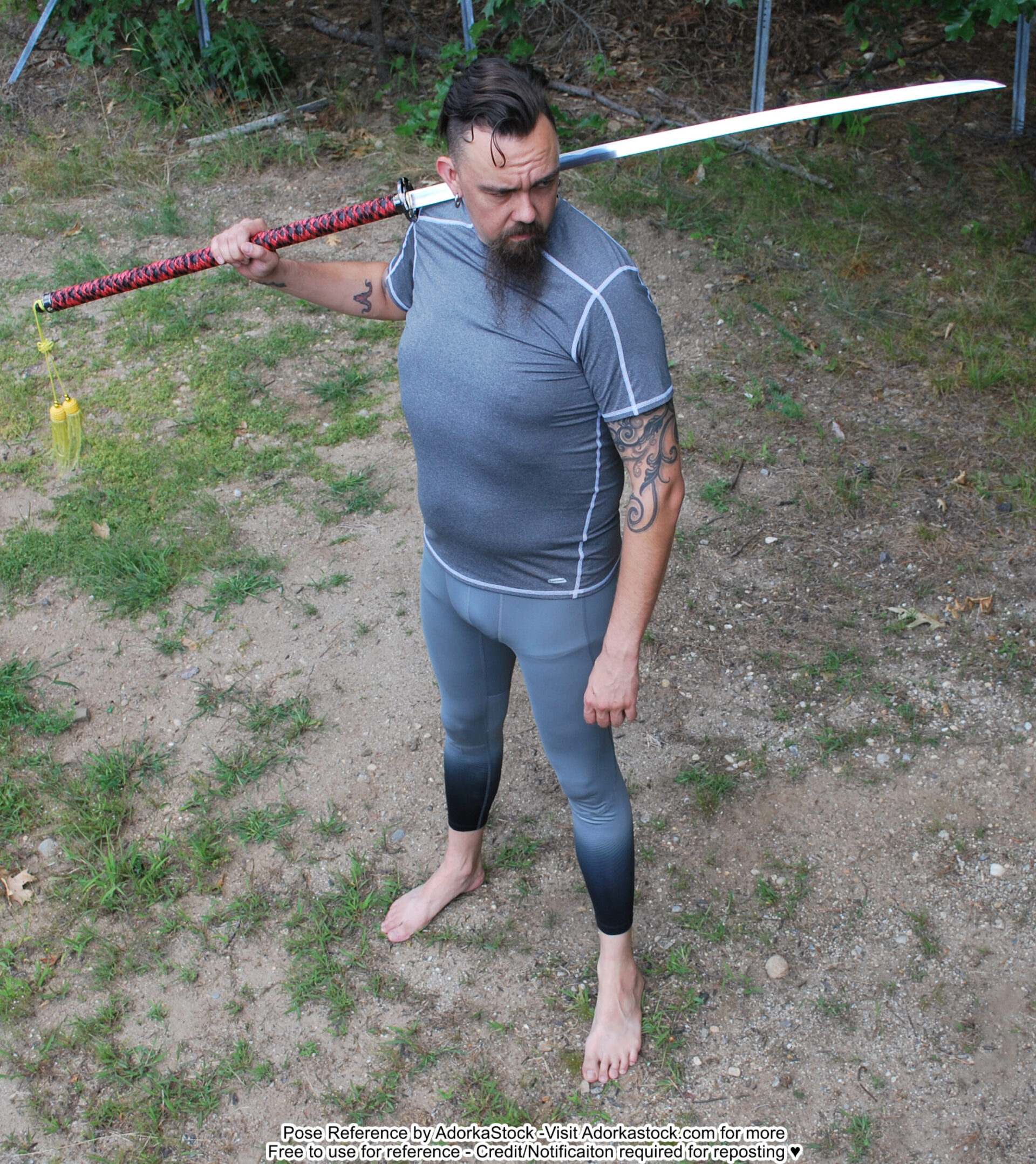 a man standing in the woods holding a katana over his shoulder. he is wearing grey pants and a grey shirt with tattoos. he is looking at something off camera.