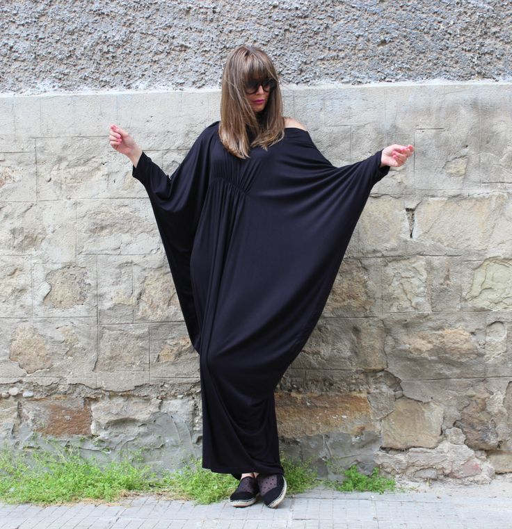 a woman with long hair and sunglasses wearing a black, oversized dress is standing in front of a stone wall. the woman has her arms outstretched.