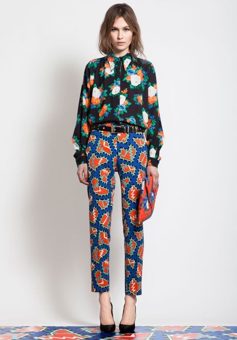 a fullbody shot of a fashion model showcasing a stylish outfit.  the model is a woman with shoulderlength light brown hair, wearing a black floral blouse with a high neckline, and vibrant blue and orange patterned cropped trousers.  she is accessorized with a black belt and a small, similarly patterned clutch.  she wears black high heels. the background is plain white. the overall style is contemporary and chic.