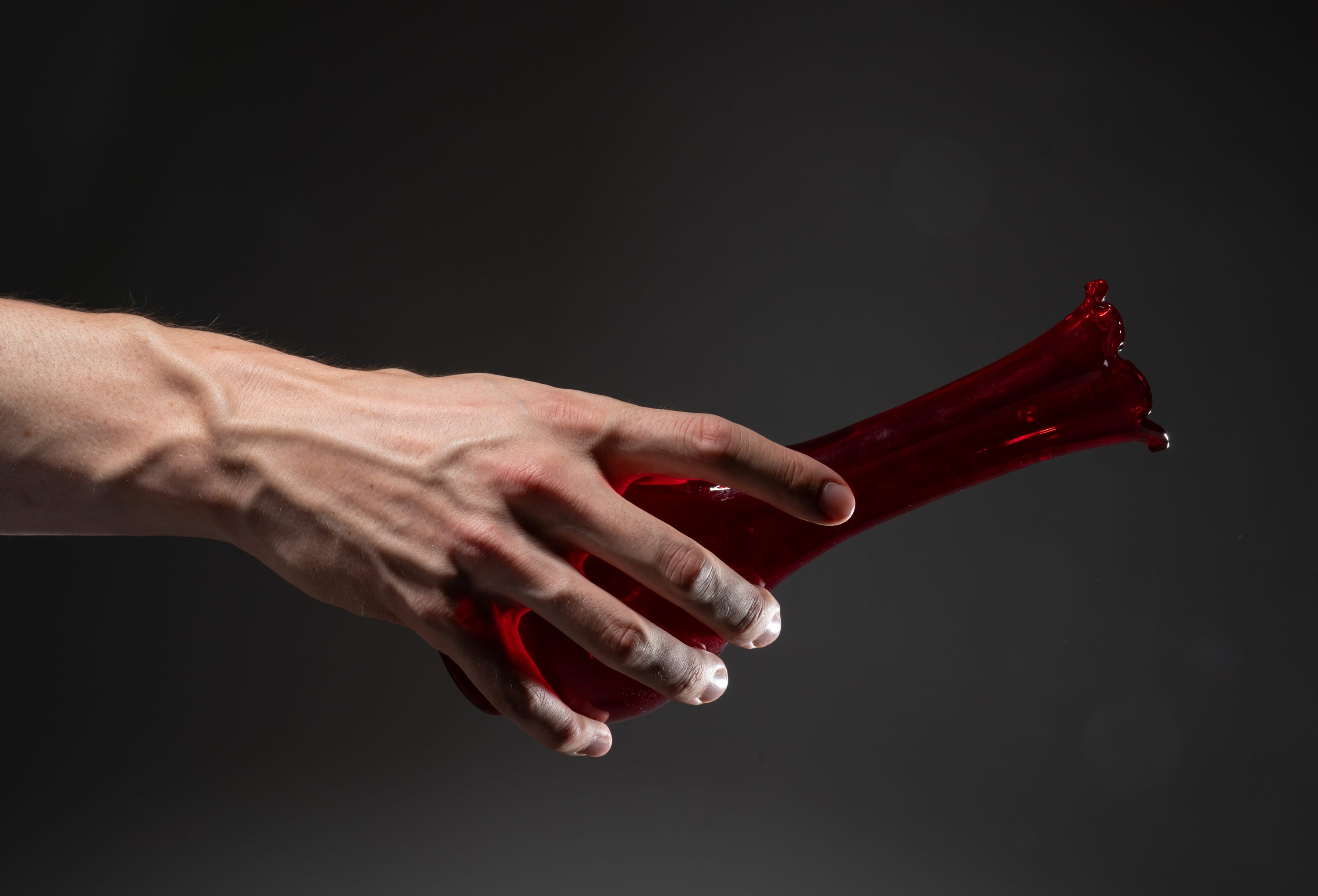 a man's hand is shown holding the broken neck of a red glass vase. the vase appears to be recently broken, with sharp edges and shards of glass still attached.  the background is a dark gray.