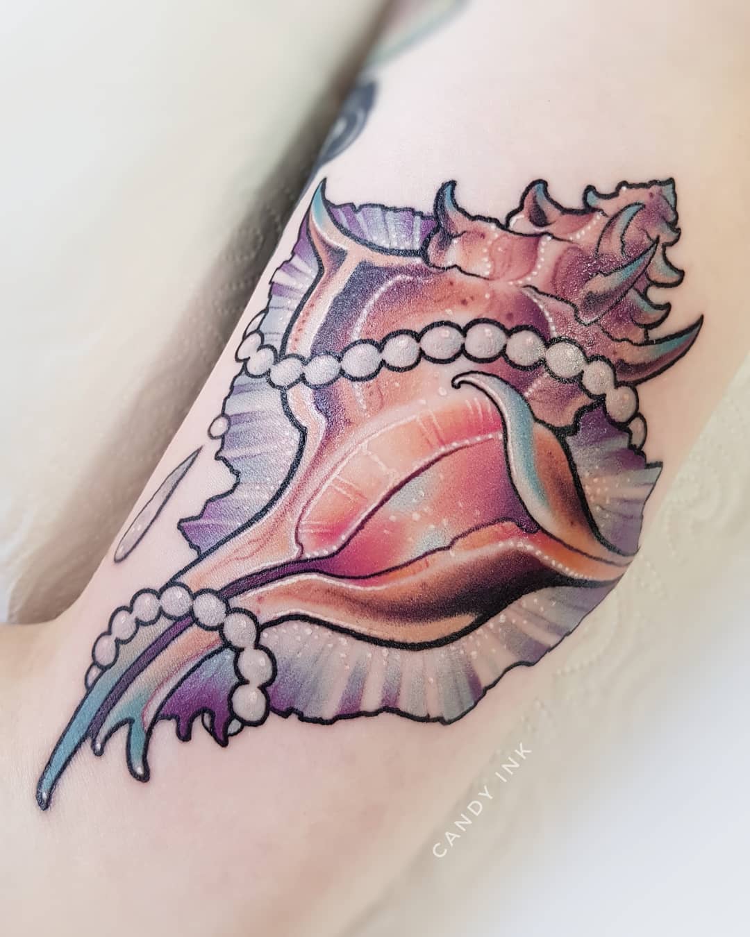 a closeup photograph showcasing a vibrant tattoo of a seashell adorned with pearls, inked on the inner forearm. the tattoo, rendered in a realistic style, boasts a blend of pink, purple, and blue hues, capturing the iridescent quality of the shell. a delicate string of pearls drapes gracefully around the shell, adding an element of elegance to the design.  the tattoo artist's signature, "candy ink," is discreetly placed below the artwork.
