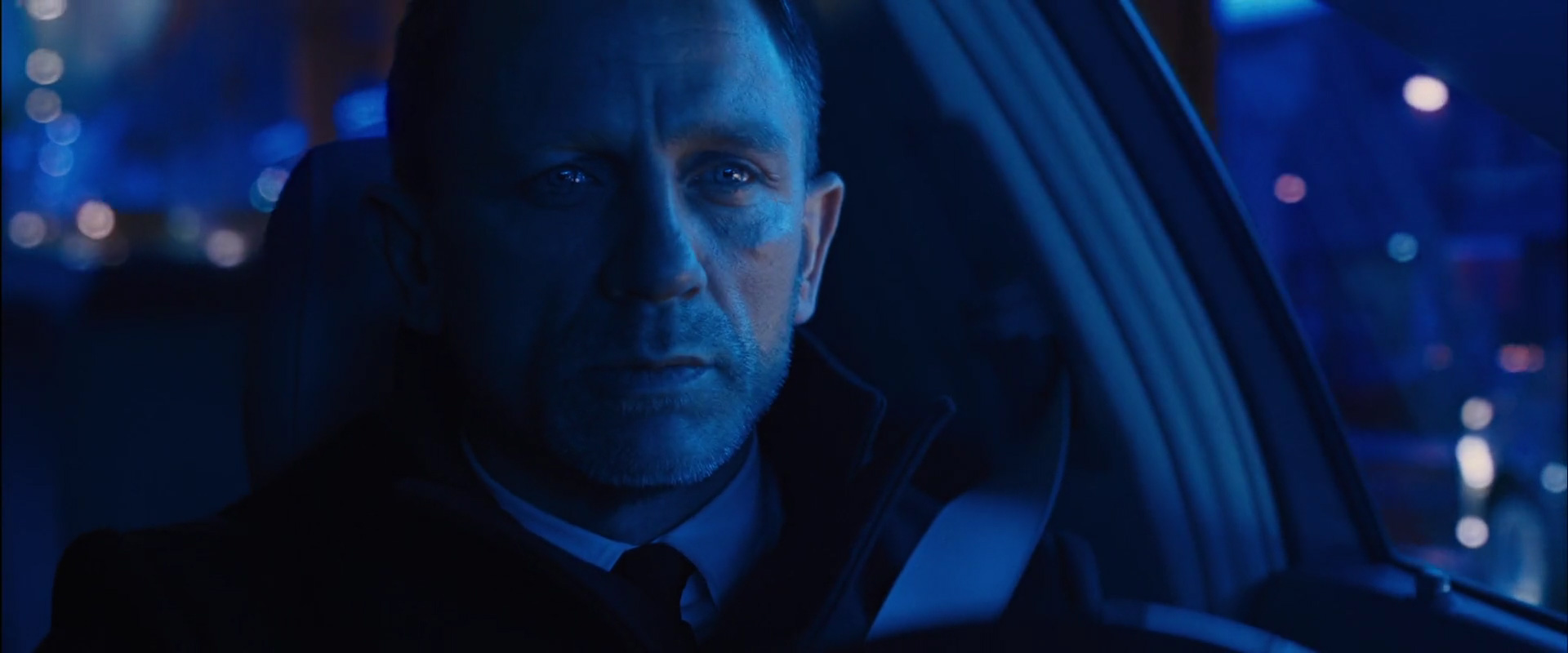 a close up shot of daniel craig as james bond in the car, looking to the side as he is driving at night. the blue light from the city and street lights shines on his face. the focus of the image is on bond's face and the intense expression in his eyes. the image captures a sense of mystery, intrigue, and danger, reflecting the themes of the spy genre.