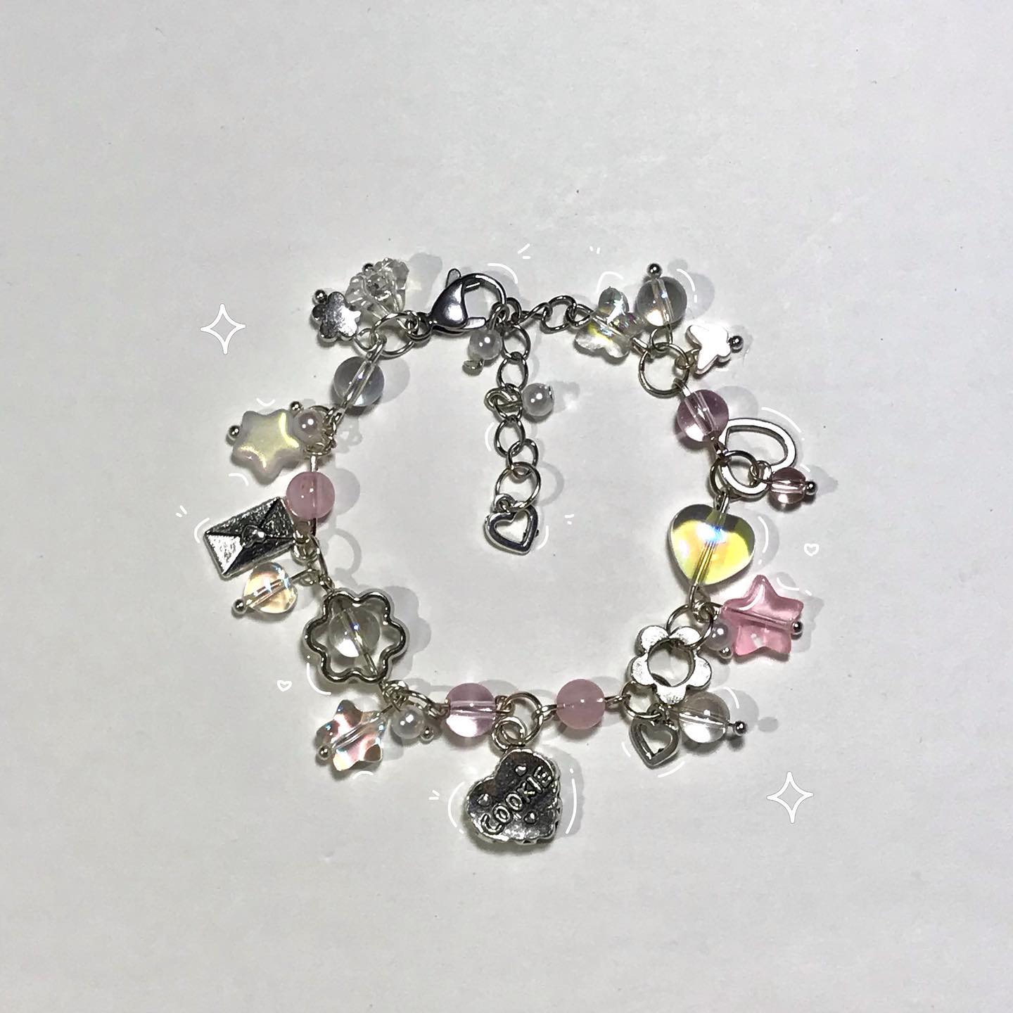 a closeup of a silver charm bracelet adorned with various charms and beads, including a "cookie" charm, flowers, hearts, stars, and clear beads. the bracelet appears to be handcrafted and has a delicate, dainty aesthetic.