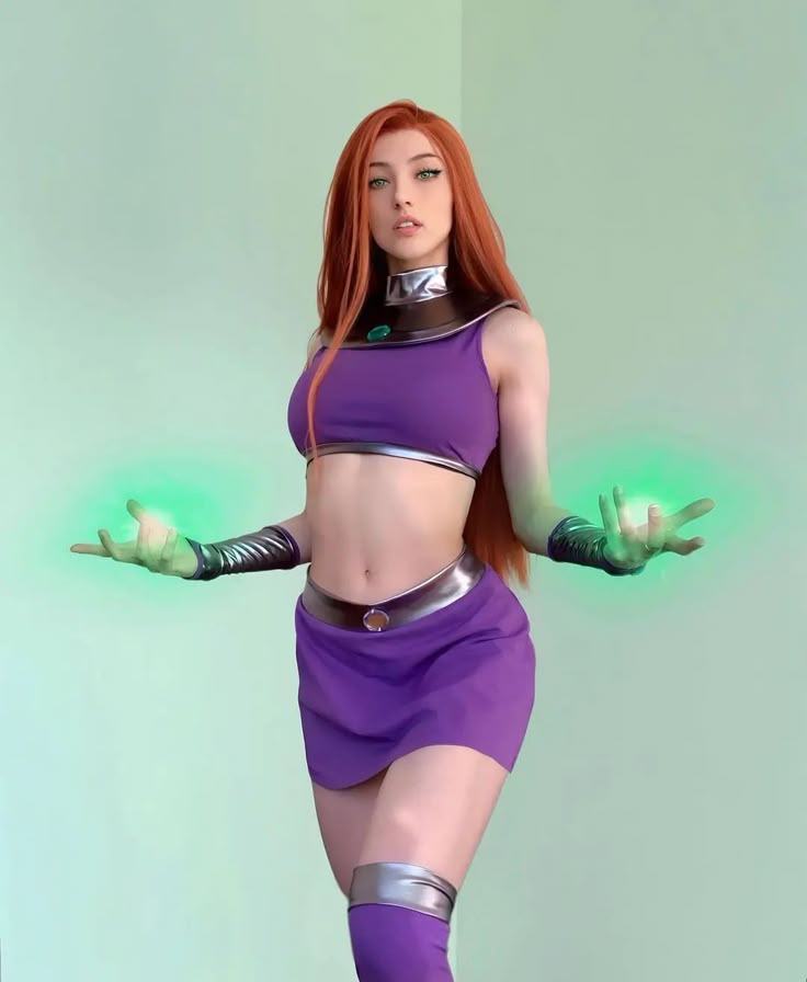 a woman dressed as starfire from dc comics' teen titans stands in a studio with her hands outstretched, glowing with green energy. she is wearing a purple crop top, a matching skirt, thighhigh socks, and silver accents on her collar, waist, gloves, and thighs. her hair is long and orange, and her eyes are green. the background is a plain light green wall.