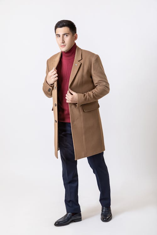 a fullbody studio shot of a stylish man wearing a camelcolored overcoat, a burgundy turtleneck sweater, dark blue trousers, and black loafers. he is standing against a white background, posing with his hands adjusting his coat. the image is welllit and showcases the man's elegant and sophisticated attire. the focus is sharp, and the clothing details are clearly visible.