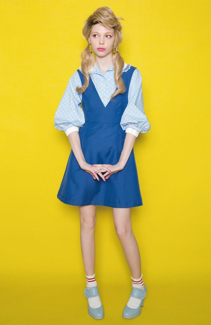 a fullbody shot of a young woman with blonde pigtails wearing a blue dress and polka dot shirt against a bright yellow background.  she's wearing light blue shoes and short white socks with red stripes. her pose is slightly demure with her hands clasped in front of her.  the overall style evokes a retro or vintage aesthetic, reminiscent of the 1960s or 1970s.