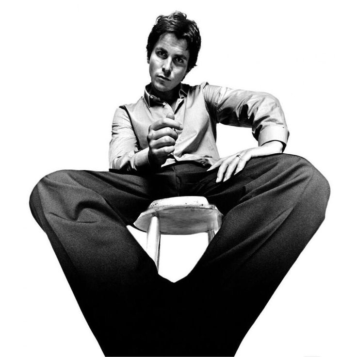 a man in a buttondown shirt and black pants sits on a stool, posing for a photo. he looks at the camera with a serious expression. the photo is shot from a low angle, creating a dramatic effect. the background is white, creating a simple and minimalist aesthetic.