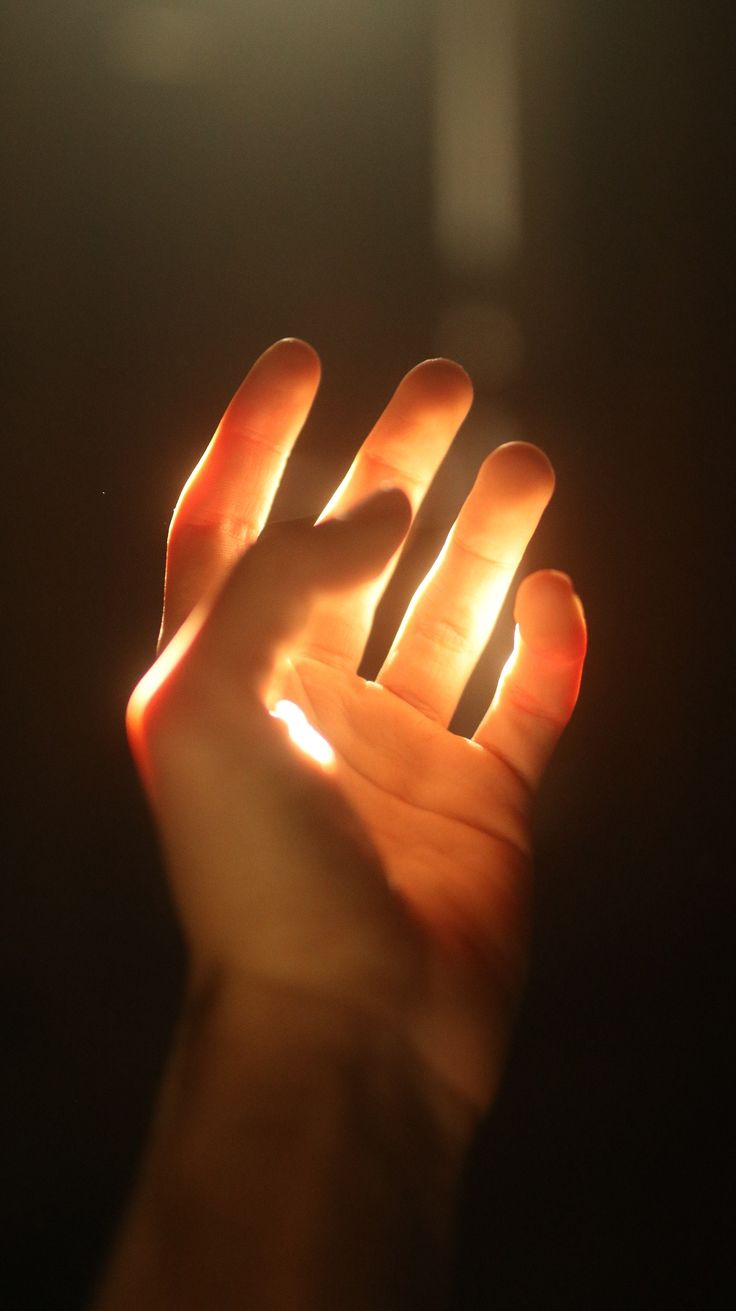 the image showcases a hand reaching up towards a source of warm, artificial light. the light illuminates the hand from below, creating a stark contrast between the illuminated fingers and the shadowed palm. the background is dark and undefined, focusing the viewer's attention on the interplay of light and shadow on the hand.